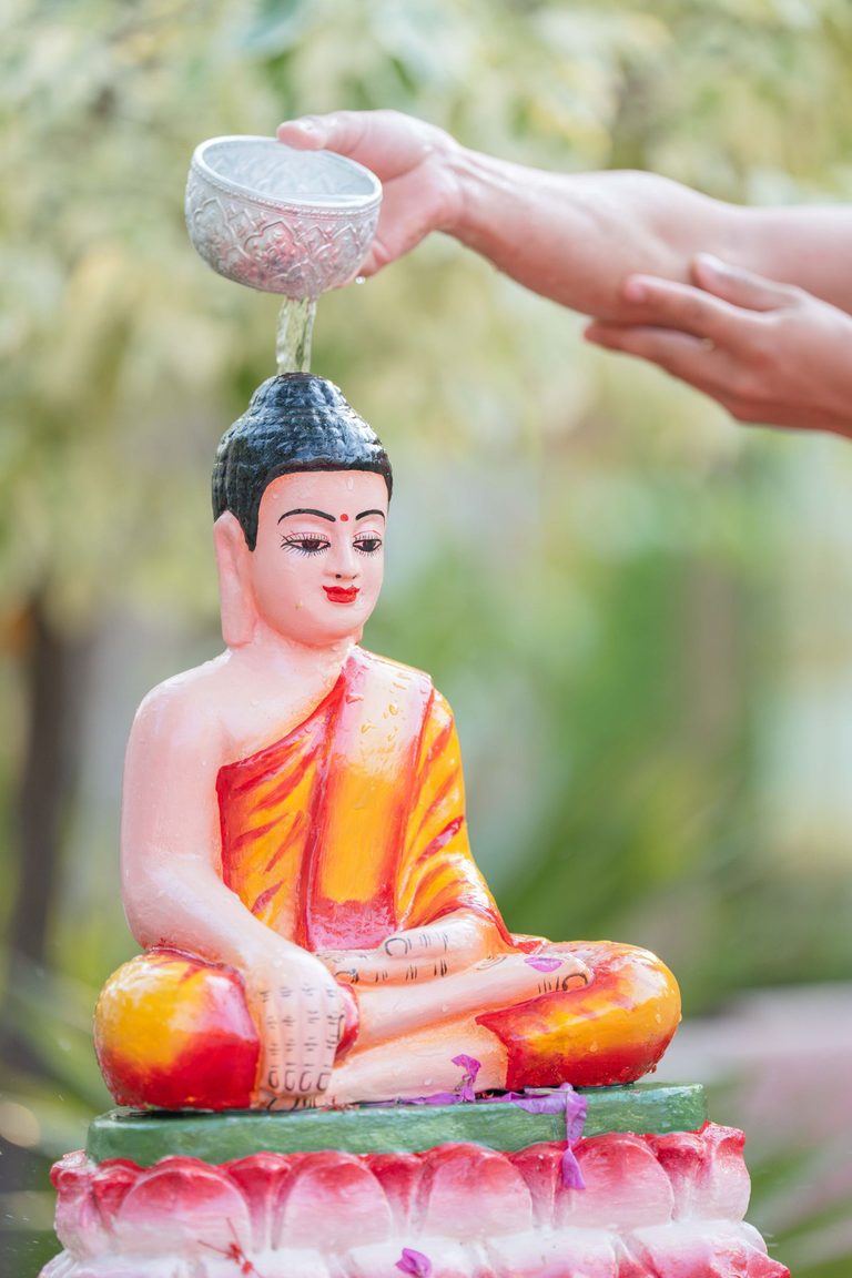Hands pour lustral water from an ornate silver bowl over a small Buddha image.