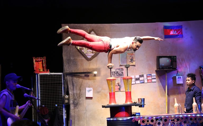 A Phare acrobat balances in a one-handed handstand on a pole under stage lights.